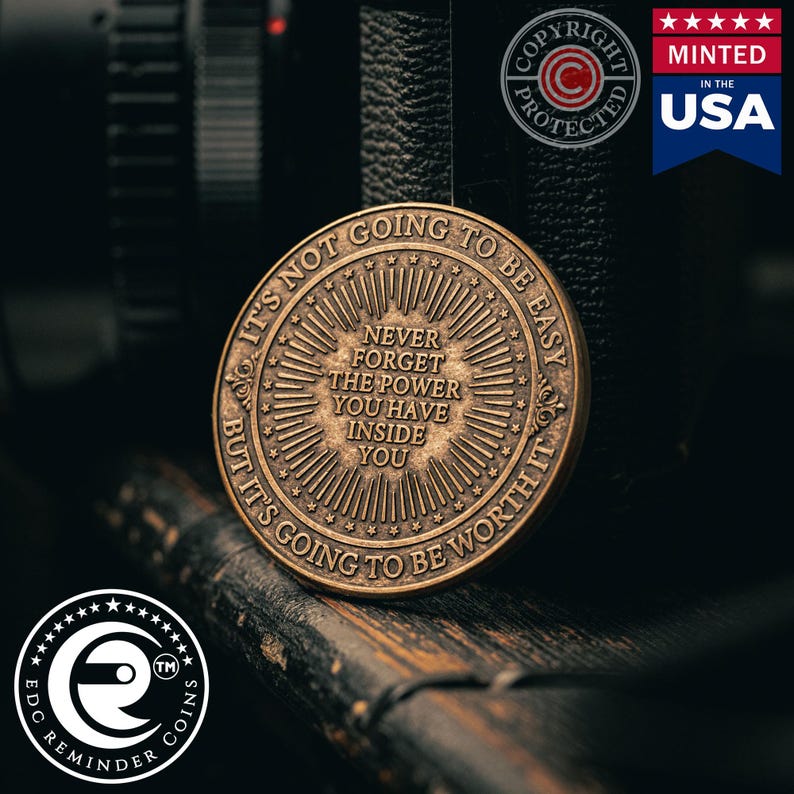 May include: A bronze-coloured coin with the inscription "It's not going to be easy, but it's going to be worth it." The coin also features the words "Never forget the power you have inside you."