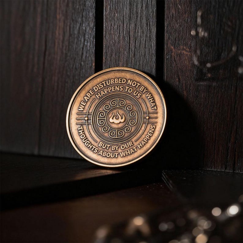 May include: A circular bronze-toned metal coin featuring an embossed central spiral design. The text WE ARE DISTURBED NOT BY WHAT HAPPENS TO US BUT BY OUR THOUGHTS ABOUT WHAT HAPPENS is engraved in a circular pattern around the center motif.