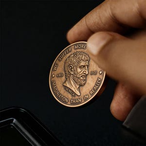 May include: A round bronze-toned metal coin featuring a relief portrait of Seneca. The text reads WE SUFFER MORE IN IMAGINATION THAN IN REALITY, with 4 BCE and 65 CE flanking the portrait. The name SENECA is inscribed below the bust.