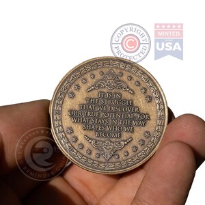 May include: A round bronze-toned metal coin featuring an embossed border of stars and laurel leaves. The center text reads: IT IS IN THE STRUGGLE THAT WE DISCOVER OUR TRUE POTENTIAL, FOR WHAT STAYS IN THE WAY SHAPES WHO WE BECOME. Decorative flourishes frame the text.