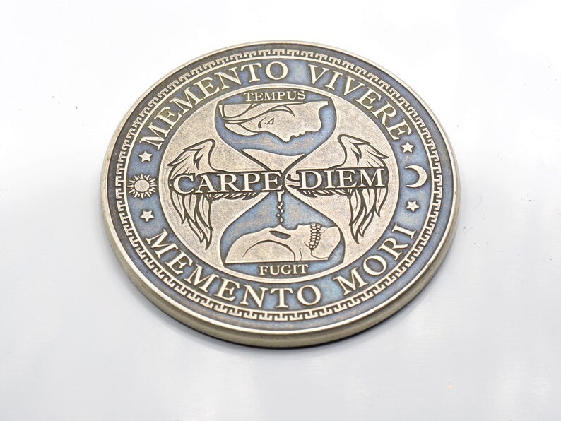 May include: A silver-toned coin with a blue background and gold lettering. The coin features an hourglass with wings and the text "Memento Vivere", "Tempus Fugit", "Carpe Diem", and "Memento Mori".