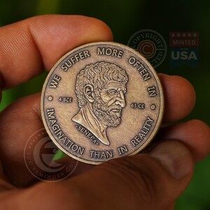 May include: A round bronze-toned metal coin featuring a relief portrait of Seneca in the center. The text reads WE SUFFER MORE OFTEN IN IMAGINATION THAN IN REALITY, with 4 BCE and 65 CE on either side of the portrait and -SENECA- below it.