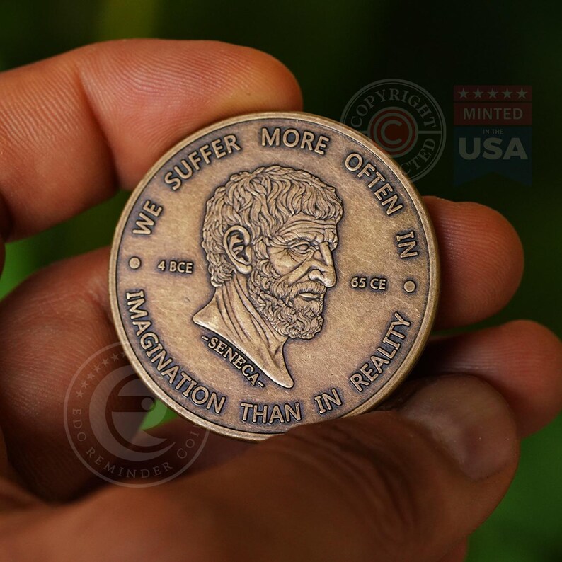 May include: A round bronze-toned metal coin featuring a relief portrait of Seneca in the center. The text reads WE SUFFER MORE OFTEN IN IMAGINATION THAN IN REALITY, with 4 BCE and 65 CE on either side of the portrait and -SENECA- below it.