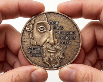 May include: A circular bronze-toned coin featuring a relief portrait of Marcus Aurelius. The text reads: Your life is what your thoughts make it, Marcus Aurelius, AD 121-180. The background of the coin has a cracked, weathered texture.