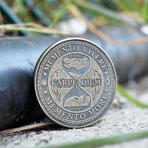 May include: A round, bronze-colored coin with an hourglass design and the Latin phrases "Memento Vivere", "Carpe Diem", and "Memento Mori".