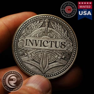 May include: A silver-toned round coin with a detailed design featuring a sword, a banner with the word "INVICTUS" and the text "BLOODY BUT UNBOWED".
