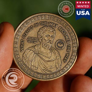 May include: A round bronze-toned metal coin featuring a relief portrait of Marcus Aurelius. The text reads "WHAT STANDS IN THE WAY BECOMES THE WAY", "MARCUS AURELIUS AD 121-180", and "STOIC" with a small flame icon, all encircled by a laurel wreath border.