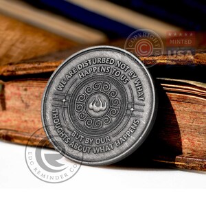 May include: A silver-toned metal coin with a quote about being disturbed by thoughts, not by what happens. The quote reads "We are disturbed not by what happens to us, but by our thoughts about what happens". The coin has a stylized flame design in the center.