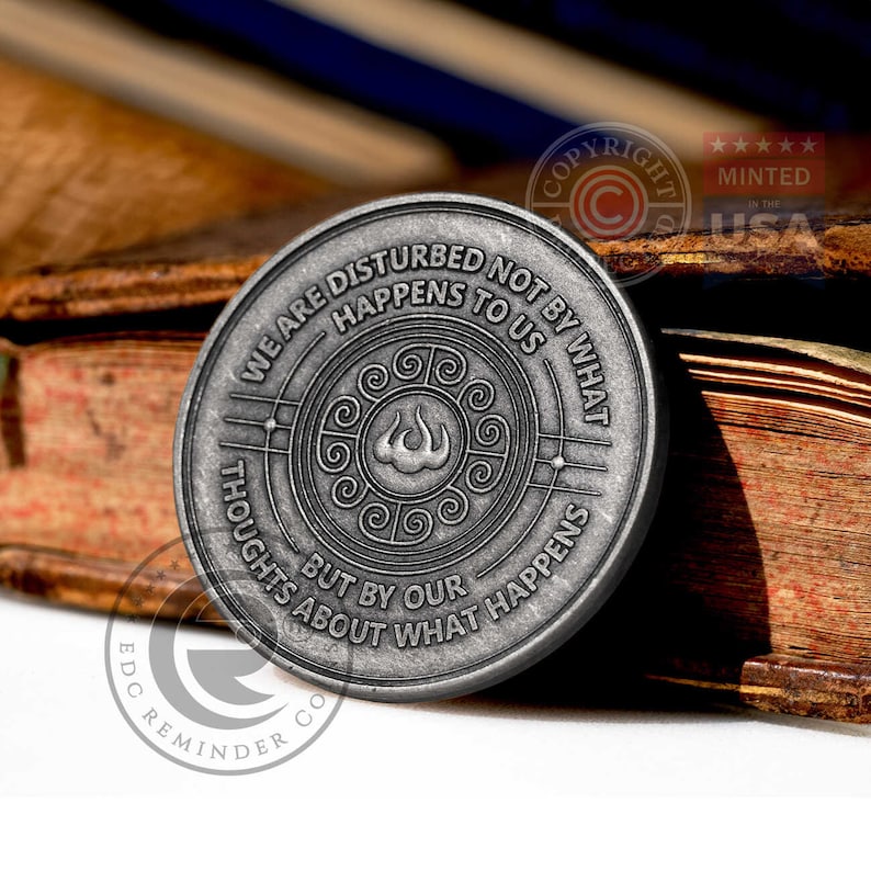 May include: A silver-toned metal coin with a quote about being disturbed by thoughts, not by what happens. The quote reads "We are disturbed not by what happens to us, but by our thoughts about what happens". The coin has a stylized flame design in the center.