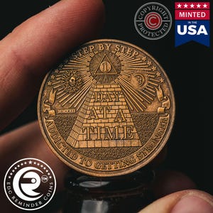 May include: A gold-toned metal coin with the words "Step by Step" and "Day at a Time" inscribed around a pyramid with an all-seeing eye at the top. The coin also has the words "Addicted to Getting Stronger" inscribed around the bottom.
