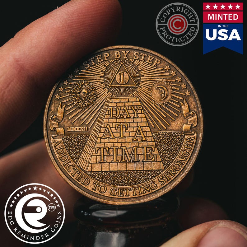 May include: A gold-toned metal coin with the words "Step by Step" and "Day at a Time" inscribed around a pyramid with an all-seeing eye at the top. The coin also has the words "Addicted to Getting Stronger" inscribed around the bottom.