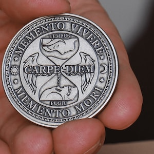 May include: A circular silver-toned metal coin featuring an hourglass design in the center with the text CARPE DIEM. The border includes the phrases MEMENTO VIVERE and MEMENTO MORI, along with the words TEMPUS and FUGIT and small star, sun, and moon icons.