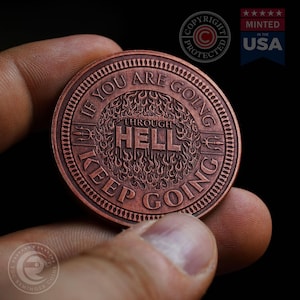 以下が含まれることがあります： 炎のグラフィックと「If you are going through hell keep going」というテキストが刻まれた銅色のコイン。コインは手で持たれている。