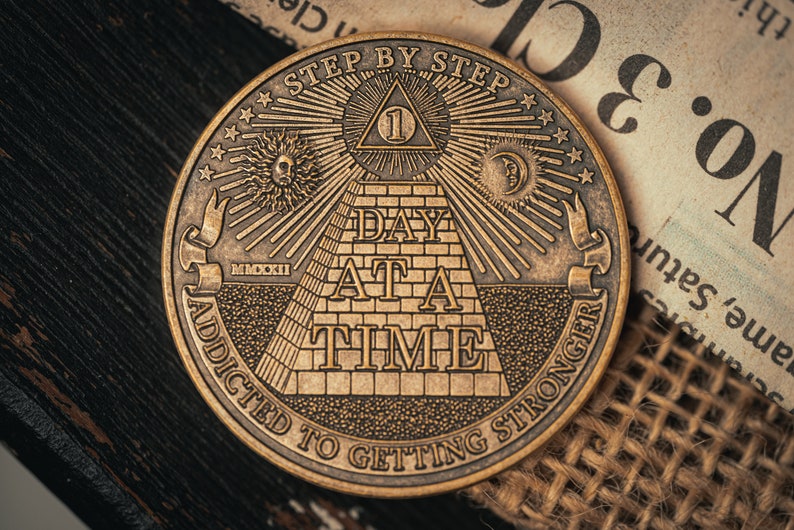 May include: A gold-toned metal coin featuring an image of a pyramid with the text "Step by Step Day at a Time Addicted to Getting Stronger". The pyramid has an eye at the top and the number "1" inside a triangle.