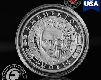 May include: A silver coin with a detailed design featuring a half-face and half-skull, surrounded by the words "MEMENTO MORI". The coin has Roman numerals and laurel leaves. The coin is "Minted in the USA".