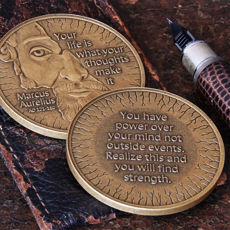 May include: Two gold-toned round metal tokens with inspirational quotes from Marcus Aurelius. The first token features a portrait of Marcus Aurelius and the quote "Your life is what your thoughts make it". The second token features the quote "You have power over your mind not outside events. Realize this and you will find strength."