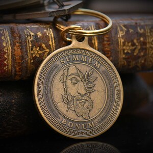 May include: A gold-toned keychain with a round, engraved medallion featuring a stylized image of a face with a laurel wreath and the text "SUMMUM BONUM".