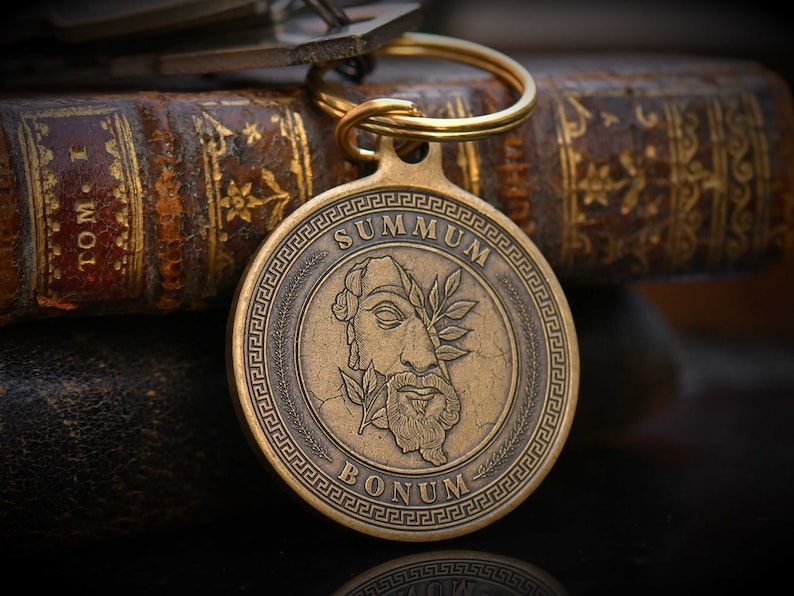 May include: A gold-toned keychain with a round, engraved medallion featuring a stylized image of a face with a laurel wreath and the text "SUMMUM BONUM".
