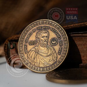 May include: A round bronze-toned metal coin featuring a relief portrait of Marcus Aurelius. The text reads "WHAT STANDS IN THE WAY BECOMES THE WAY" around the edge, with "MARCUS AURELIUS AD 121-180" and a small flame icon inside a laurel wreath.