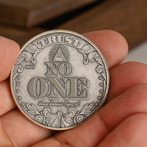 May include: A silver-toned coin with the words "TRUST NO ONE" and an all-seeing eye symbol. The coin also has the year "MMXX" and the Latin phrase "Homo homini lupus est" inscribed on it.