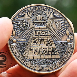 May include: A gold-toned metal coin with an all-seeing eye pyramid design. The coin features the text "Step by Step" and "Addicted to Getting Stronger" around the edge. The centre of the coin features the text "Day at a Time".