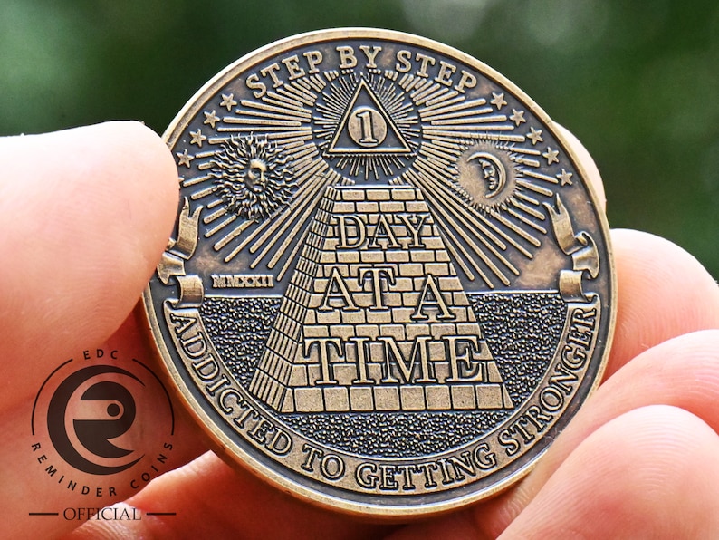 May include: A gold-toned metal coin with an all-seeing eye pyramid design. The coin features the text "Step by Step" and "Addicted to Getting Stronger" around the edge. The centre of the coin features the text "Day at a Time".