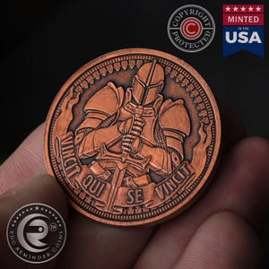 May include: A copper-colored coin with a knight in armor holding a sword. The coin has the words "VINCIT QUI SE VINCIT" around the edge.