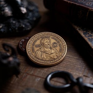 May include: A circular bronze-toned metal coin featuring a relief portrait of Marcus Aurelius. The text "WHAT STANDS IN THE WAY BECOMES THE WAY" is embossed around the border, along with the name "MARCUS AURELIUS" and a small flame icon.