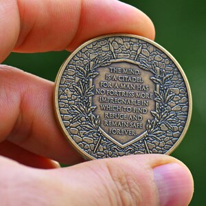 Op de afbeelding: Een ronde, bronskleurige metalen munt met een verhoogd schildontwerp in het midden met de tekst: THE MIND IS A CITADEL, FOR A MAN HAS NO FORTRESS MORE IMPREGNABLE IN WHICH TO FIND REFUGE AND REMAIN SAFE FOREVER, omringd door laurierbladeren.