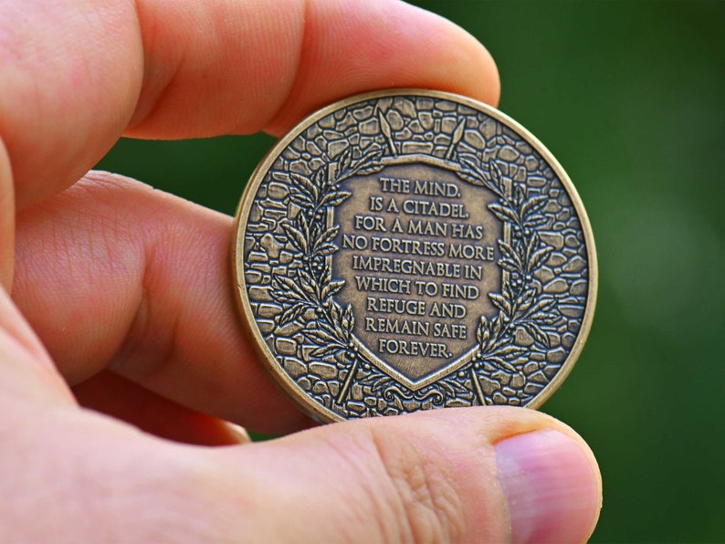 Op de afbeelding: Een ronde, bronskleurige metalen munt met een verhoogd schildontwerp in het midden met de tekst: THE MIND IS A CITADEL, FOR A MAN HAS NO FORTRESS MORE IMPREGNABLE IN WHICH TO FIND REFUGE AND REMAIN SAFE FOREVER, omringd door laurierbladeren.