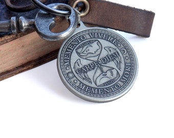 May include: A round silver-toned metal medallion keychain featuring an hourglass design with wings, a skull, and the text MEMENTO VIVERE, CARPE DIEM, MEMENTO MORI, TEMPUS, and FUGIT. The medallion is attached to a metal ring and a brown leather strap.