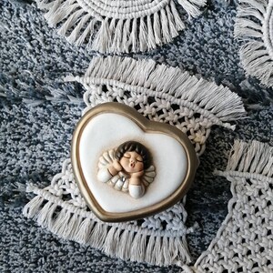 May include: A white ceramic heart-shaped trinket box with a gold trim. The box features a relief sculpture of a sleeping angel with a crown and a blue gem on its chest. The box is sitting on a white crocheted doily with fringe.