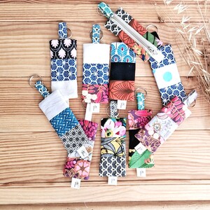 May include: A collection of fabric keychains with various patterns and colors, including floral, geometric, and abstract designs. Each keychain has a metal ring and a small tag. The keychains are arranged on a wooden surface.