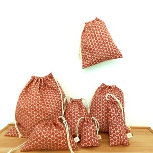 May include: Set of six red drawstring bags with a white geometric pattern. The bags are of various sizes and are arranged on a wooden surface.