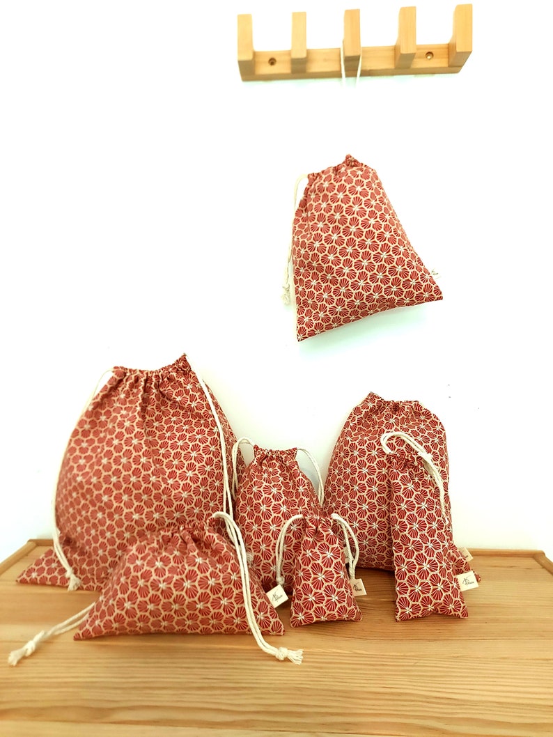 May include: Set of six red drawstring bags with a white geometric pattern. The bags are of various sizes and are arranged on a wooden surface.