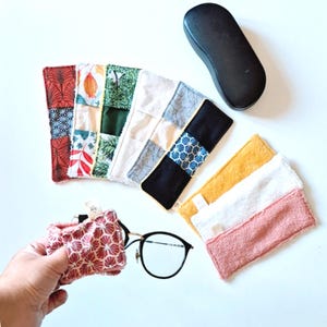Könnte beinhalten: Eine Sammlung farbenfroher, gemusterter Brillenetuis und Reinigungstücher. Die Etuis weisen Patchwork-Designs mit verschiedenen Stoffen und Mustern auf. Ein schwarzes Brillenetui und eine Brille sind ebenfalls vorhanden. Die Reinigungstücher sind in Gelb, Weiß und Rosa.
