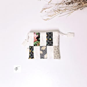 Op de afbeelding: Een witte zak met trekkoord en een patchwork-ontwerp. Het patchwork bevat bloemen-, geometrische en bladpatronen in verschillende kleuren. De zak heeft een eenvoudige, minimalistische uitstraling.