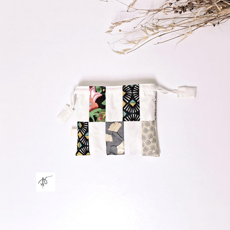 Op de afbeelding: Een witte zak met trekkoord en een patchwork-ontwerp. Het patchwork bevat bloemen-, geometrische en bladpatronen in verschillende kleuren. De zak heeft een eenvoudige, minimalistische uitstraling.