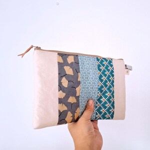 Op de afbeelding: Een rechthoekig, beige etui met ritssluiting en een patchwork-ontwerp. Het patchwork omvat delen van grijze stof met gele bladerpatronen, blauwe geometrische vormen en turquoise en witte waaiervormige ontwerpen. Een hand houdt het etui tegen een witte achtergrond.