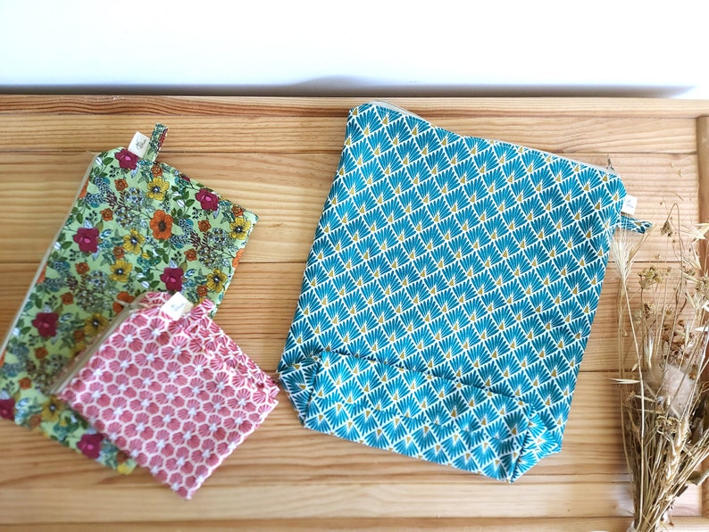 May include: Three fabric pouches with zipper closures rest on a wooden surface. One is teal with a geometric pattern, another is green with a floral print, and the third is pink with a honeycomb design. The pouches vary in size.