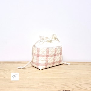 Könnte beinhalten: Ein weiß-rosa gesteppter Kordelzugbeutel mit Karomuster. Die Tasche hat einen gerafften oberen Teil und einen dicken, cremefarbenen Kordelzug. Die Tasche steht auf einer hellen Holzoberfläche.