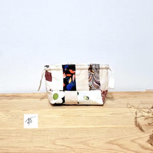 May include: A small, rectangular patchwork fabric pouch with a drawstring closure. The pouch is made of various patterned fabrics in cream, black, red, blue, and floral designs. The pouch sits on a wooden surface.