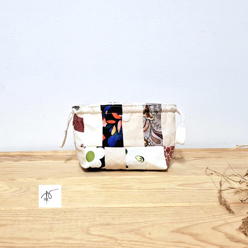 May include: A small, rectangular patchwork fabric pouch with a drawstring closure. The pouch is made of various patterned fabrics in cream, black, red, blue, and floral designs. The pouch sits on a wooden surface.
