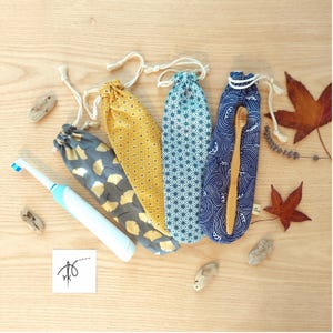 Può includere: Tre sacchetti in tessuto con coulisse e motivi vari, tra cui foglie di ginkgo dorate su grigio, forme geometriche gialle e disegni a onde blu. Uno spazzolino elettrico bianco e uno spazzolino in legno sono posizionati accanto.