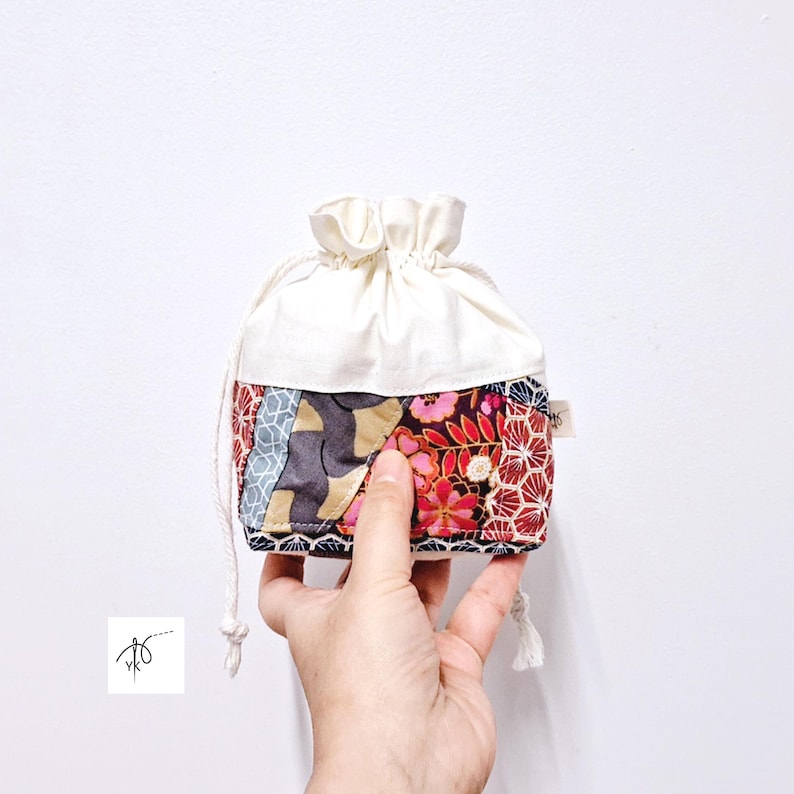May include: A small, drawstring pouch with a patchwork design. The lower portion features a mix of floral and geometric patterns in red, pink, blue, and gold. The upper part is a solid cream color. A white drawstring and tassel complete the design.