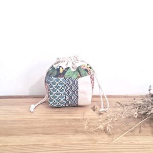 Pode incluir: Uma pequena bolsa de cordão branco com um design de patchwork que apresenta vários padrões, incluindo padrões florais, geométricos e abstratos. A bolsa é feita de tecido de algodão e tem um fecho de cordão branco.