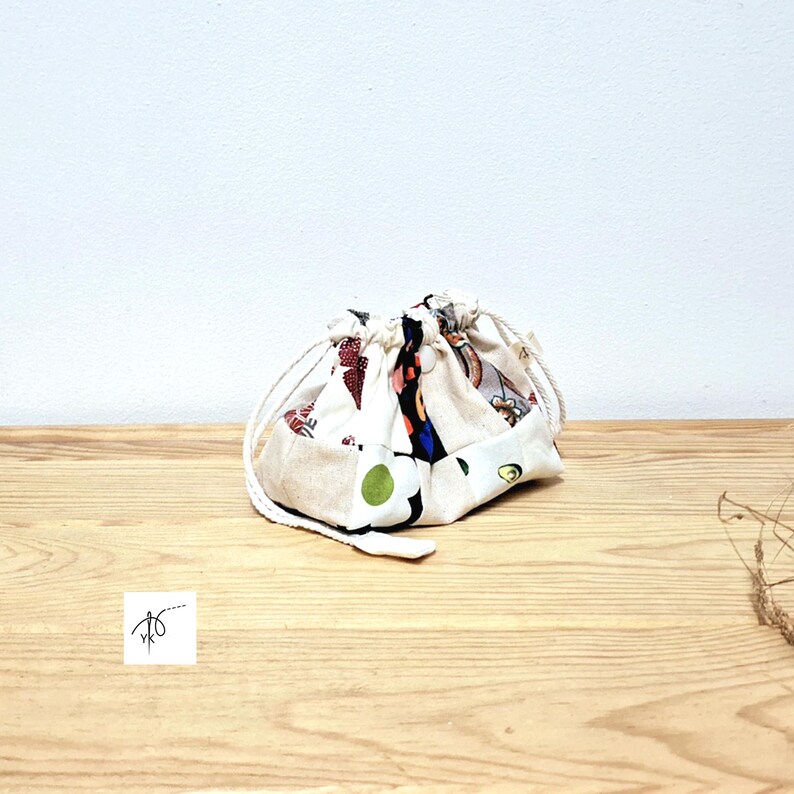 May include: A small drawstring bag with a cream background and a patchwork design featuring red, green, and black floral and geometric patterns. The bag is on a wooden surface.