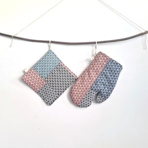 Peut inclure: Deux maniques de cuisine avec un motif géométrique dans des tons de bleu, rose et noir. Les maniques sont suspendues à une branche.