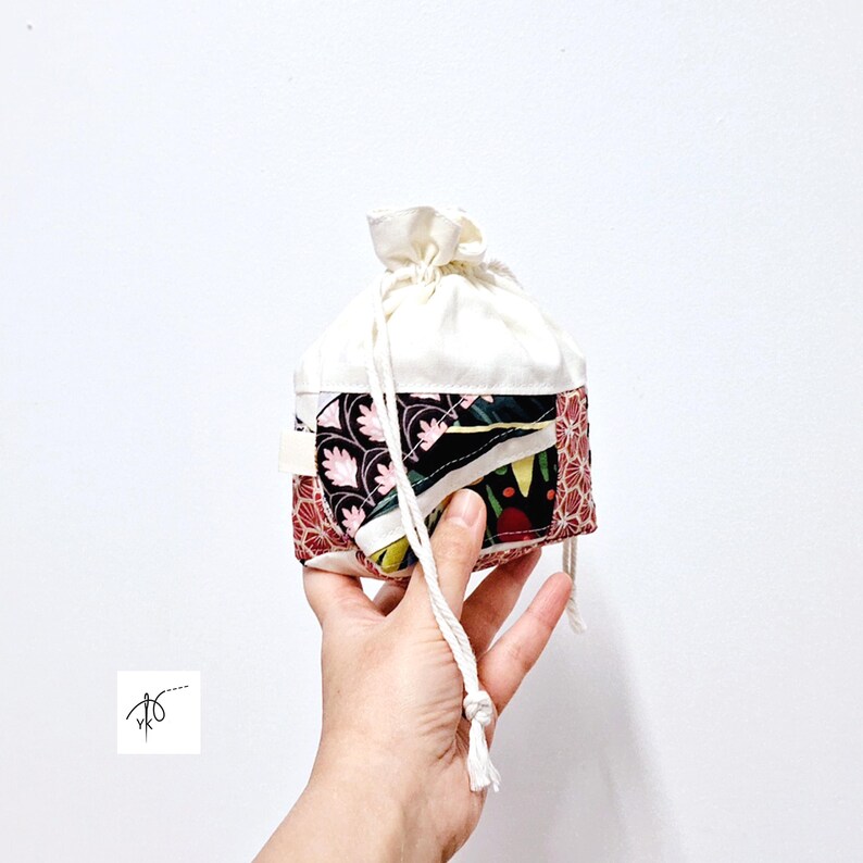 May include: A small, drawstring pouch with a patchwork design. The pouch features a cream-colored top and a patterned lower section with floral and geometric motifs in red, black, and green. A white drawstring secures the top.
