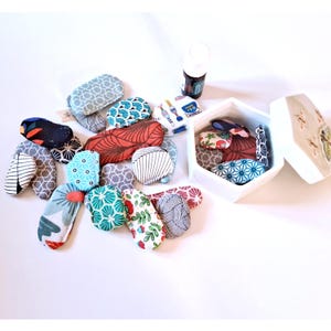 Op de afbeelding: Een verzameling kleine stoffen items met diverse patronen en kleuren, waaronder blauw, rood en grijs. Sommige zijn verspreid, andere liggen in een witte zeshoekige doos. Een flesje etherische olie is ook aanwezig.